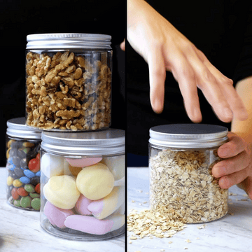 Vasetti trasparenti con tappo in alluminio pieno di biscotti ed adesivo prodotto artigianale retto tra due mani altri cibi